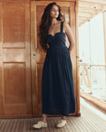 A woman with long hair wears THE GREAT. SU26 Day Break Dress in navy and beige shoes, standing on a wooden deck before closed doors.