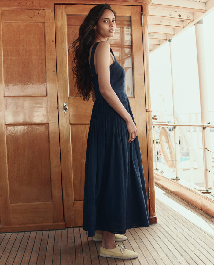 A woman in THE GREAT. SU26 The Day Break Dress in navy and light shoes stands on a wooden deck before a wooden door, glancing over her shoulder.