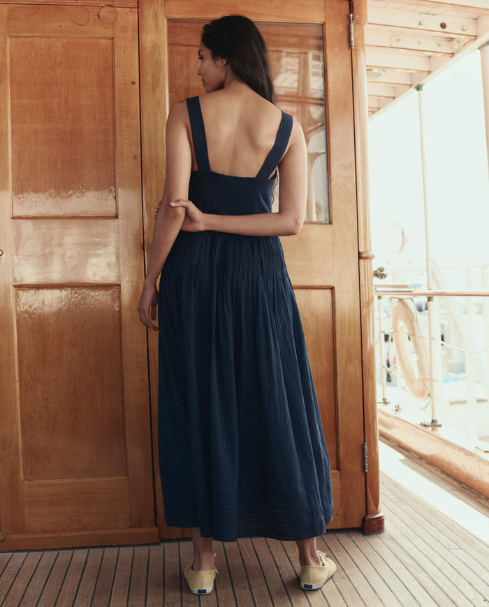 A woman stands on a wooden deck facing away, wearing THE GREAT. SU26 The Day Break Dress in navy with sandals, her left arm relaxed at her side.