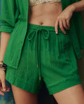 Wearing THE GREAT. SP26 SWIM's The Delta Short in Bright Sprig, paired with a bright green button-up, patterned top underneath, and bracelets on the left wrist.