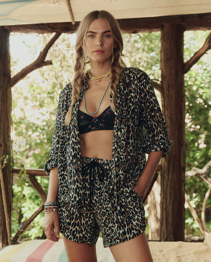 A woman with braided hair stands indoors by large windows in lush greenery, wearing THE GREAT. SP26 SWIM's The Delta Short in leopard print over a black floral bikini top.