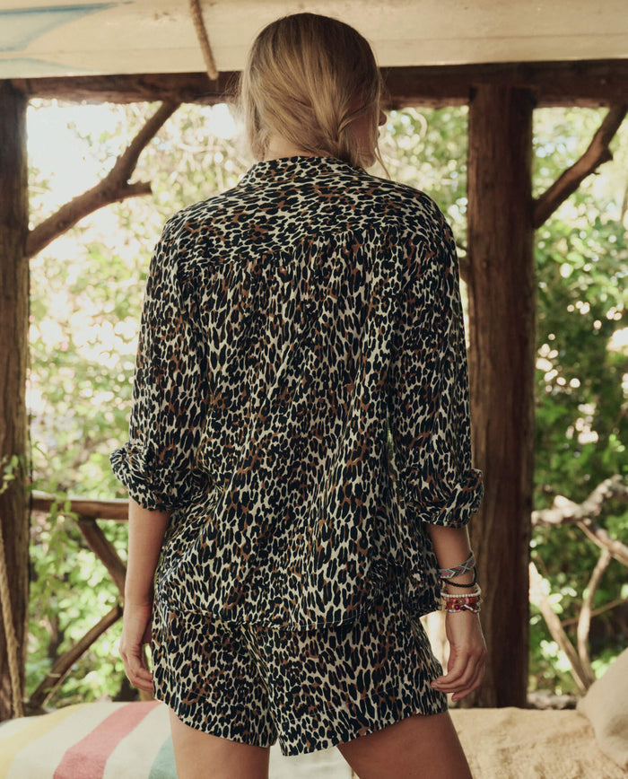 A person stands with their back to the camera, wearing THE GREAT. SP26 SWIM’s The Delta Short in leopard, surrounded by trees and wooden beams in the background.