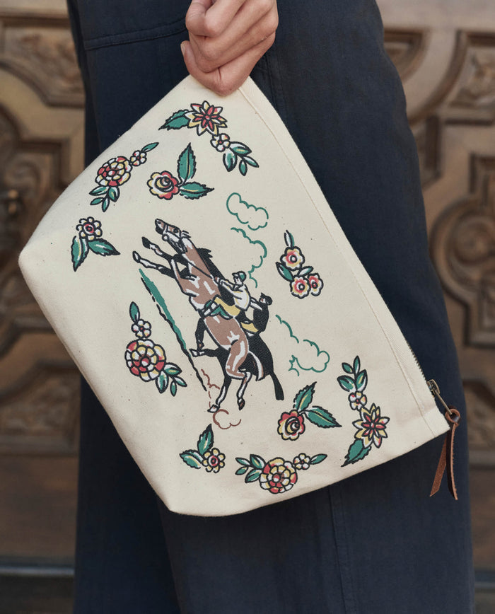 A person holds The Derby Pouch in cream by THE GREAT. PF26, featuring an embroidered horse and rider with floral designs.