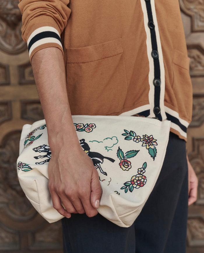 A person in a brown sweater holds THE GREAT. PF26 The Derby Pouch in cream, featuring floral and horse designs.