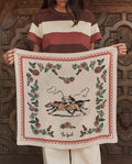 A person holds THE GREAT. PF26 The Derby Silk Bandana in cream, featuring a central illustration of two horse riders with floral motifs and a geometric border.