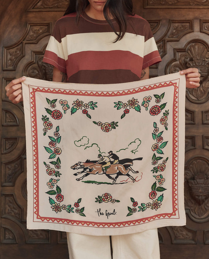 A person holds THE GREAT. PF26 The Derby Silk Bandana in cream, featuring a central illustration of two horse riders with floral motifs and a geometric border.