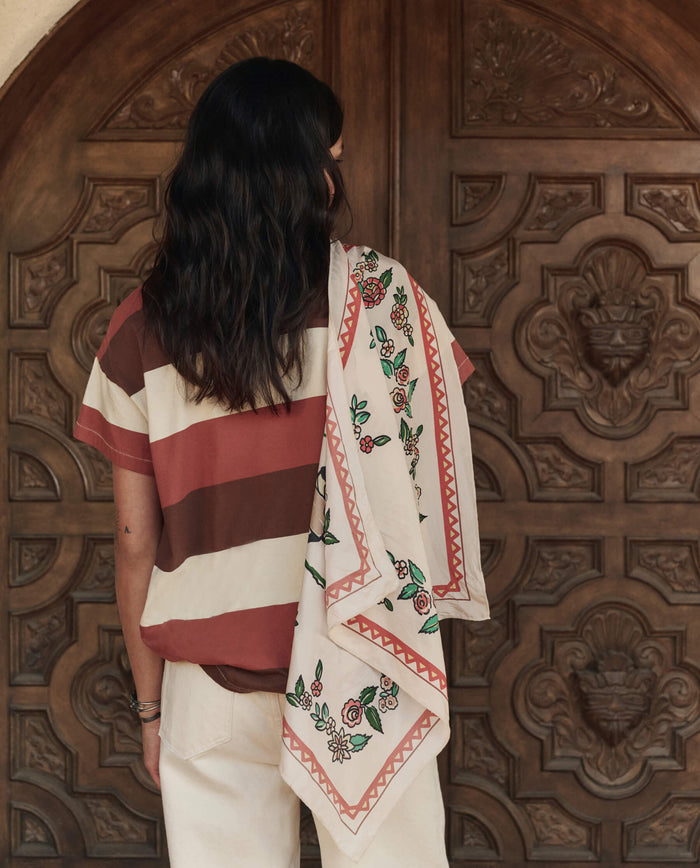 A person with long dark hair faces a carved wooden door, wearing a striped shirt and light pants, with THE GREAT. PF26's The Derby Silk Bandana in Cream draped stylishly over their shoulder.