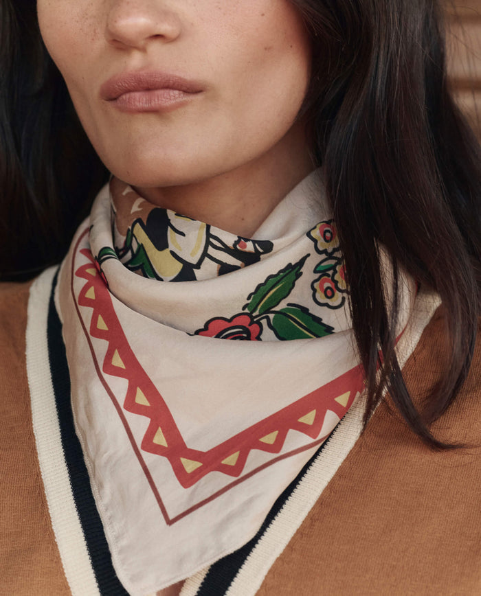 A woman wears a tan sweater with black and white trim and The Derby Silk Bandana in cream by THE GREAT. PF26, featuring red, green, yellow, and black designs around her neck.