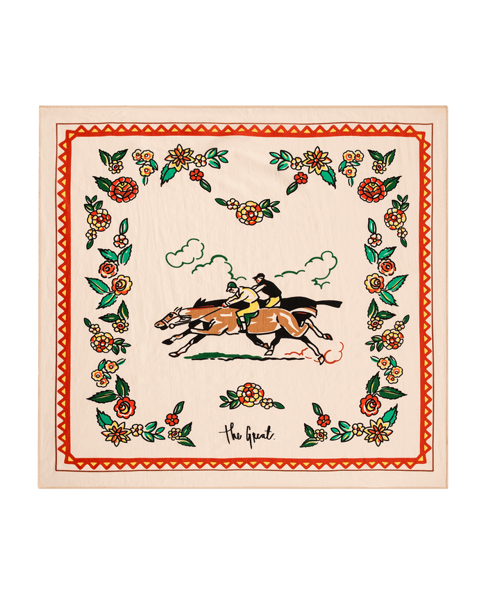 The Derby Silk Bandana by THE GREAT. PF26—Cream, showcases two jockeys racing horses at the center, framed by a floral border and “The Great.” at the bottom, making it a stylish accessory for the Derby.
