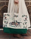 A person in a cream dress holds THE GREAT. PF26 The Derby Tote – Cream, featuring horse racing and floral designs, while standing on tiled flooring before a wooden door.