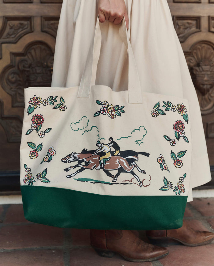 A person in a cream dress holds THE GREAT. PF26 The Derby Tote – Cream, featuring horse racing and floral designs, while standing on tiled flooring before a wooden door.