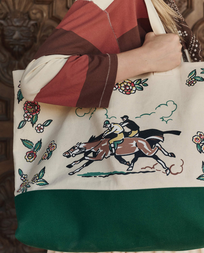A person holds The Derby Tote in Cream by THE GREAT. PF26, featuring horse racing illustrations and floral patterns, paired with a striped long-sleeve top.