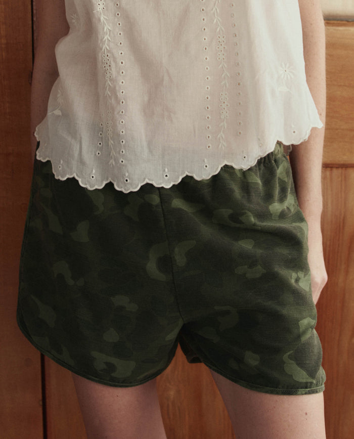 A person wearing a white eyelet blouse and THE GREAT. SU26 The Dolphin Short in Desert Camo stands in front of a wooden door.