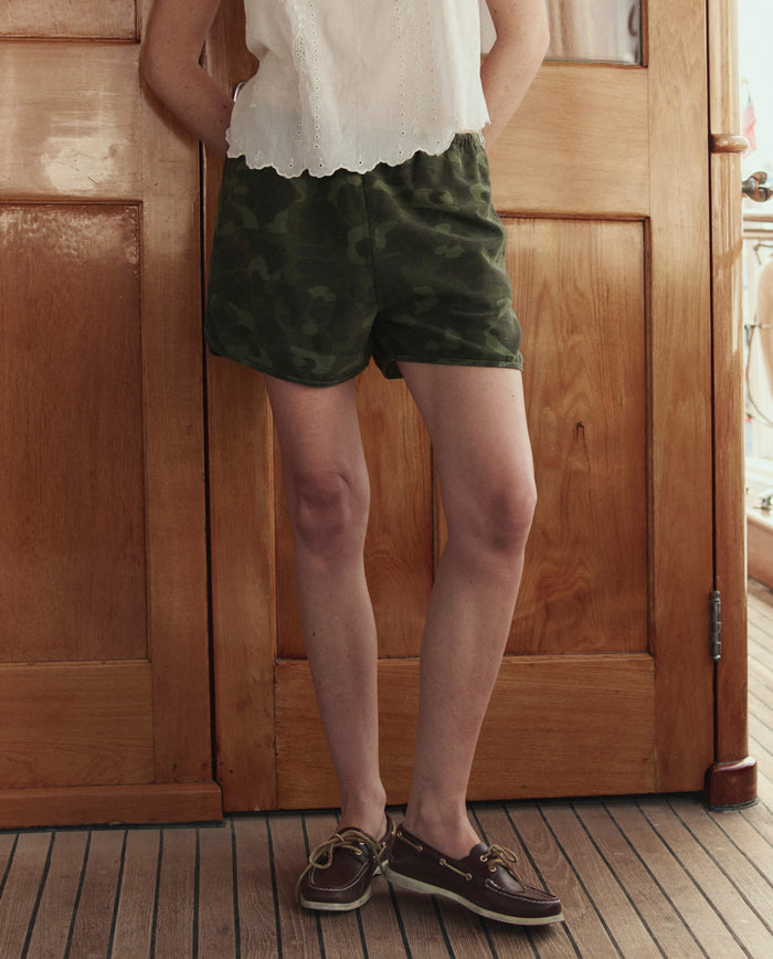 A person stands indoors before wooden doors, wearing a white sleeveless top, THE GREAT. SU26 The Dolphin Short in Desert Camo, and brown boat shoes.