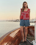 At sunset, a woman stands on a wooden boat in THE GREAT. SU26 Dolphin Short -- Shoreline Wash, a red patterned sleeveless top, and boat shoes, with sailboats and calm water in the background.