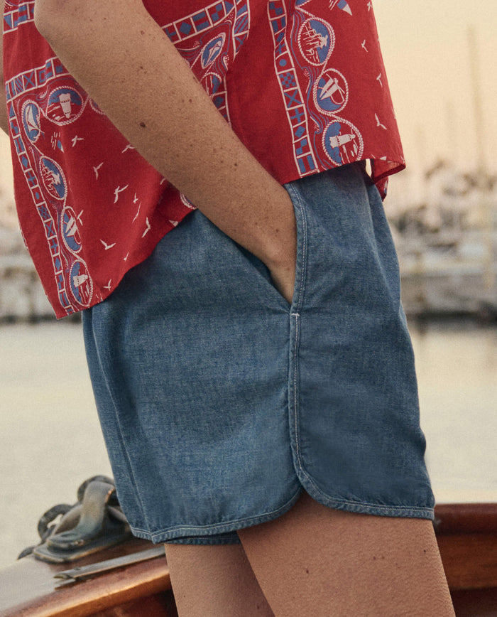 Standing on a boat with hands in the pockets of THE GREAT. SU26 The Dolphin Short -- Shoreline Wash, and wearing a red patterned shirt; marina and boats appear in the background.