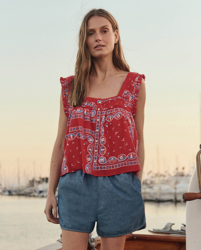 A woman stands on a boat at sunset, wearing THE GREAT. SU26’s 'The Dolphin Short. -- Shoreline Wash' and a red patterned sleeveless top, with a marina and sailboats in the background.