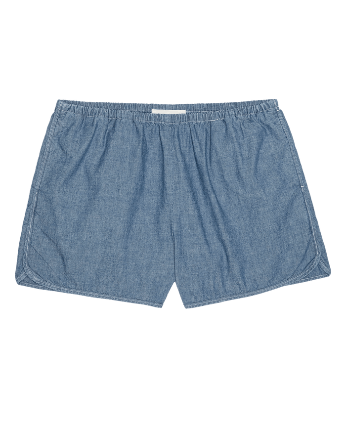 THE GREAT. SU26 The Dolphin Short in Shoreline Wash—a light blue chambray short with an elastic waistband, shown flat on a white background.