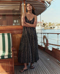 A woman stands with arms crossed on a wooden boat deck, wearing THE GREAT. SU26’s The Drop Waist Dress in Black And Cream Token Floral, with water and buildings in the background.