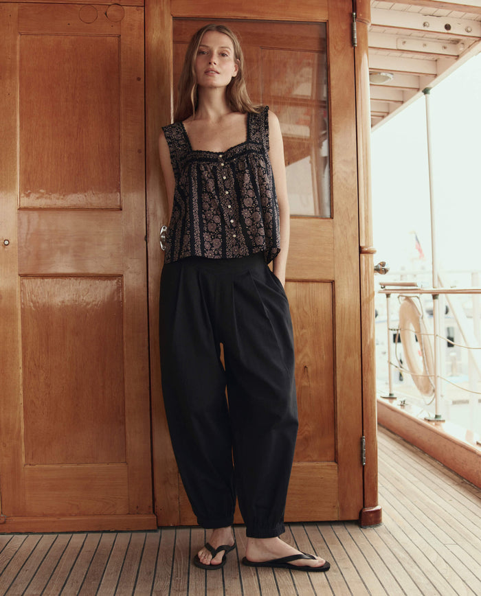 A woman stands on a wooden deck wearing THE GREAT. SU26 The Elastic Blouson Pant in black, paired with a sleeveless patterned top and black sandals, her arms relaxed by her sides.