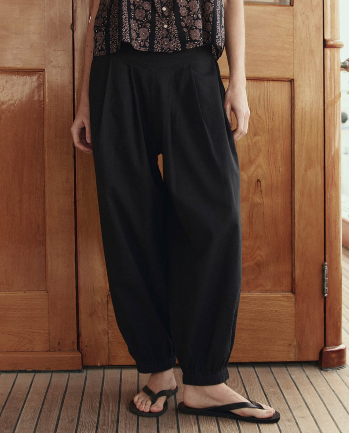 Someone stands indoors on a wood floor in front of wooden doors, wearing THE GREAT. SU26 The Elastic Blouson Pant in black, paired with a patterned top and black flip-flops.