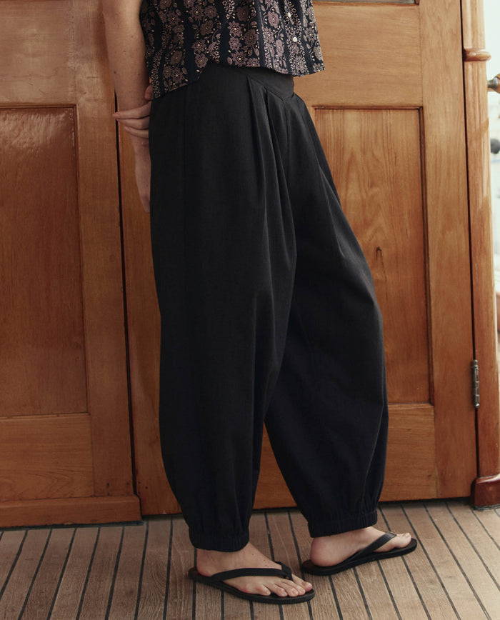 A person wearing THE GREAT. SU26 The Elastic Blouson Pant in black, a sleeveless patterned top, and black flip-flops stands indoors on a wooden floor before a wooden door.