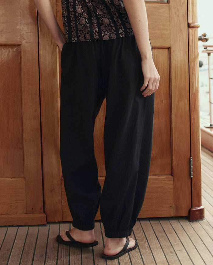 Seen from behind indoors, a person wears THE GREAT. SU26 The Elastic Blouson Pant in black, paired with a patterned top and black sandals, with one arm relaxed by their side.