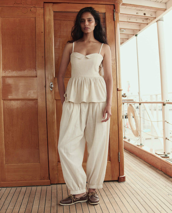 A woman stands on a wooden deck wearing THE GREAT. SU26 The Elastic Blouson Pant in Natural, paired with a sleeveless cream peplum top and brown boat shoes.