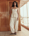 A woman stands on a wooden deck wearing THE GREAT. SU26 The Elastic Blouson Pant in Natural, paired with a sleeveless cream peplum top and brown boat shoes.