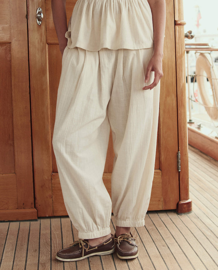 A person wearing THE GREAT. SU26 The Elastic Blouson Pant in Natural, paired with a matching top and brown boat shoes, stands on a wooden floor with wooden walls and railing in the background.