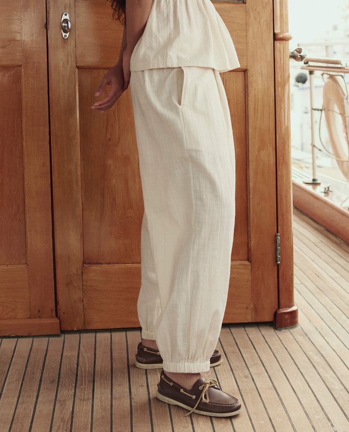 A person stands on a wooden deck wearing THE GREAT. SU26 The Elastic Blouson Pant in Natural, paired with a matching top and brown boat shoes.