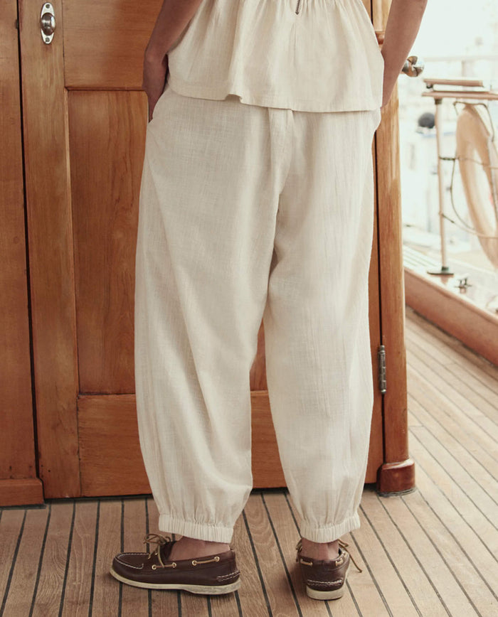 A person stands on a wooden deck, seen from behind, wearing THE GREAT. SU26 The Elastic Blouson Pant in Natural with a matching top and brown boat shoes.