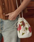 A person in light blue jeans and a brown shirt carries THE GREAT. SU26 Embroidered Cinched Bucket Bag in white with red and white floral details.