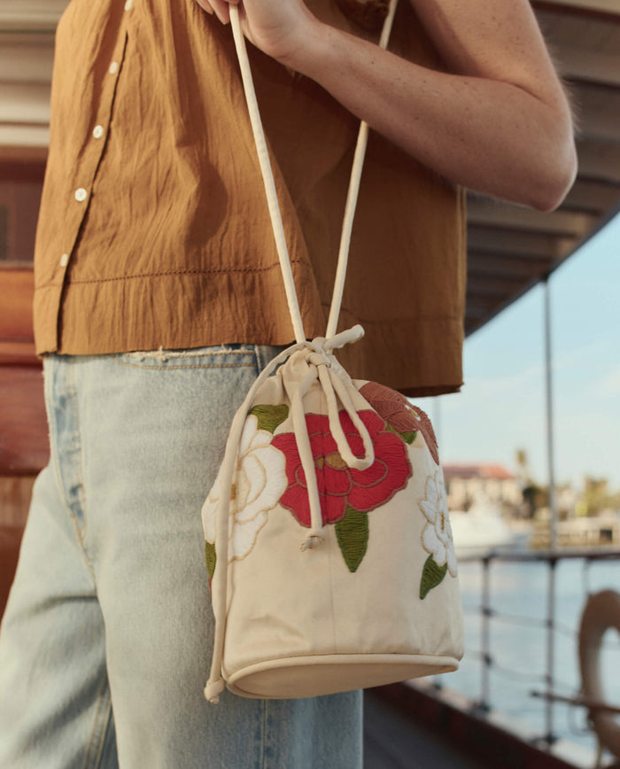 A person in light blue jeans and a brown sleeveless top holds The Embroidered Cinched Bucket Bag in white with red and white floral details by THE GREAT. SU26.