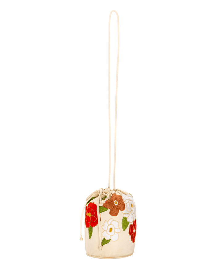 The Embroidered Cinched Bucket Bag by THE GREAT. SU26 is a small white bucket bag with red, white, and brown floral embroidery, green leaves, a long strap, and a charming drawstring closure.