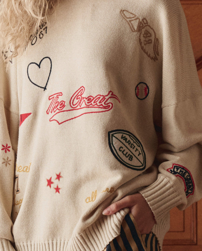 A person wearing THE GREAT. SP26 D1 The Embroidered Oversize Crew Pullover in cream, featuring patches and designs like "The Great," a heart, and a varsity club emblem.