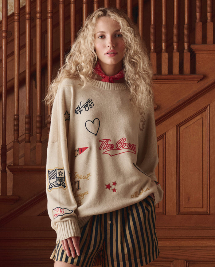 A person with curly blonde hair stands indoors in THE GREAT. SP26 D1 The Embroidered Oversize Crew Pullover in cream, in front of a wooden staircase.