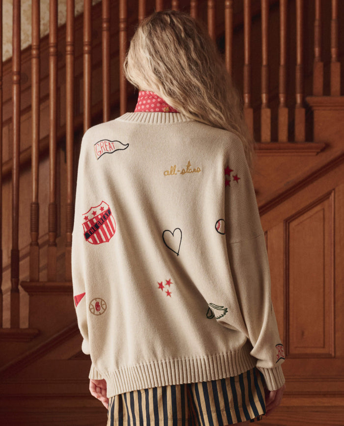 A person with long, wavy blonde hair stands in front of a wooden staircase, wearing THE GREAT. SP26 D1 The Embroidered Oversize Crew Pullover in Cream and black-and-gold striped pants.