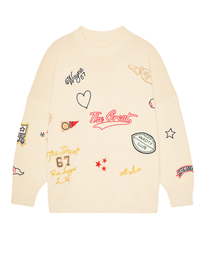 The Embroidered Oversize Crew Pullover in cream by THE GREAT. SP26 D1 features colorful patches—stars, hearts, text, and sporty designs—on a cozy, oversized crewneck sweater.