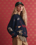 A person with long curly blonde hair wears THE GREAT. SP26 D1 The Embroidered Oversize Crew Pullover in True Navy over a striped shirt, tan pants, and a dark cap, standing against a red patterned background.
