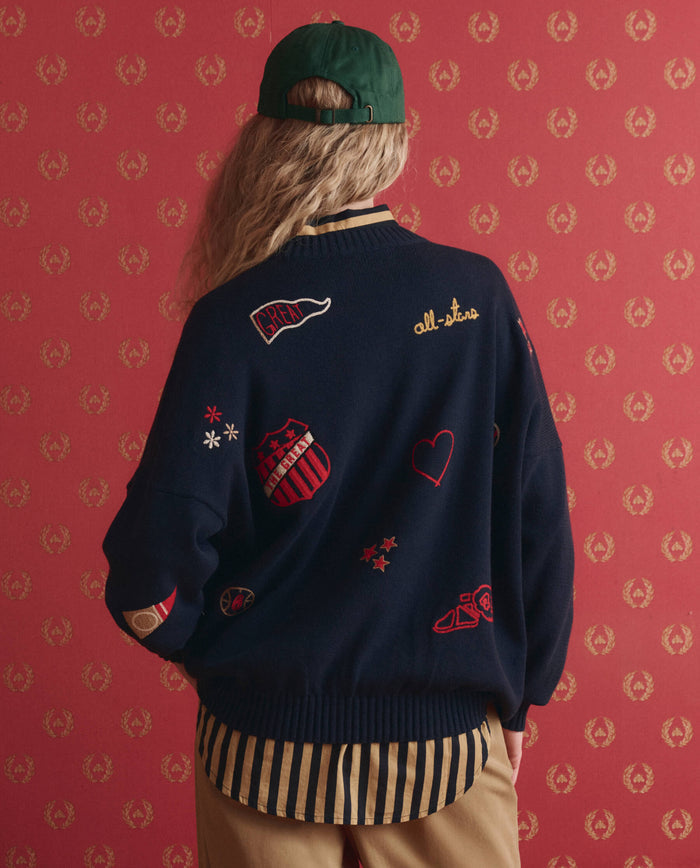 A person faces away from the camera, wearing THE GREAT. SP26 D1 The Embroidered Oversize Crew Pullover in True Navy, tan pants, and a green cap, standing before a red patterned wall.