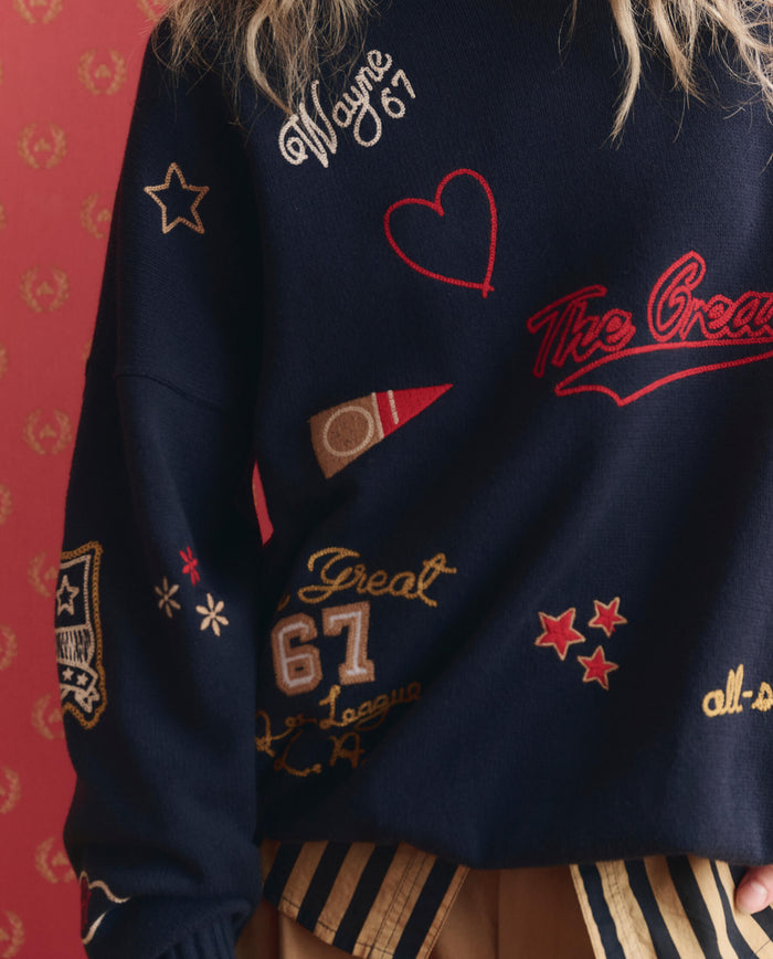 A person wears THE GREAT. SP26 D1 The Embroidered Oversize Crew Pullover in True Navy, featuring star and heart patches, numbers, and "Wayne 67" text, standing against a red patterned background.