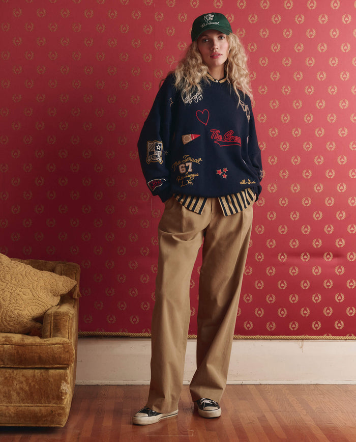 A person in THE GREAT. SP26 D1 The Embroidered Oversize Crew Pullover in True Navy, green cap, and wide tan pants stands by a gold couch against a red patterned wall.