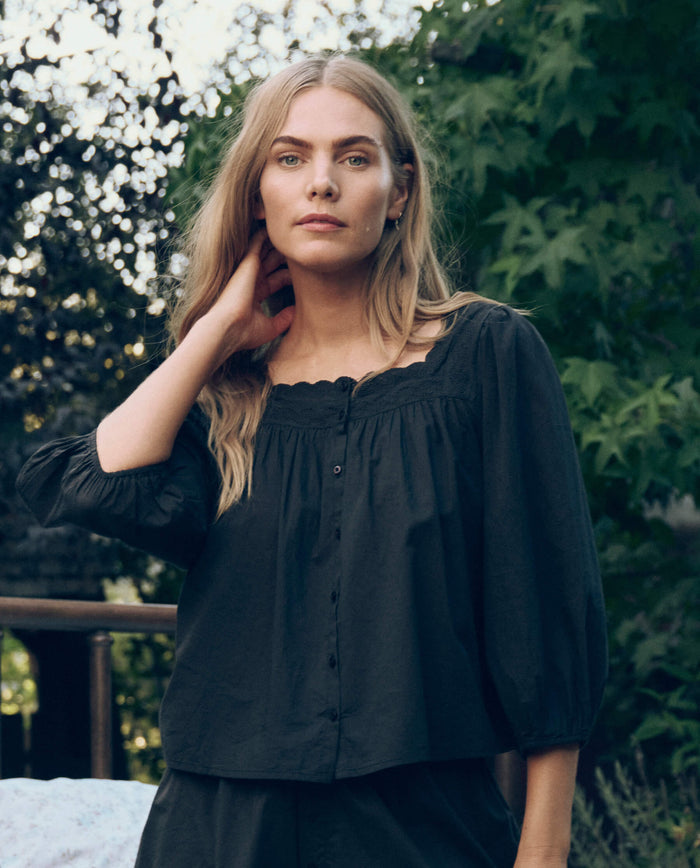 A woman with long blonde hair stands outdoors among trees, wearing THE GREAT. CORE SLEEP's The Eyelet Button Sleep Top in black, featuring three-quarter sleeves and Victorian-inspired lace details.
