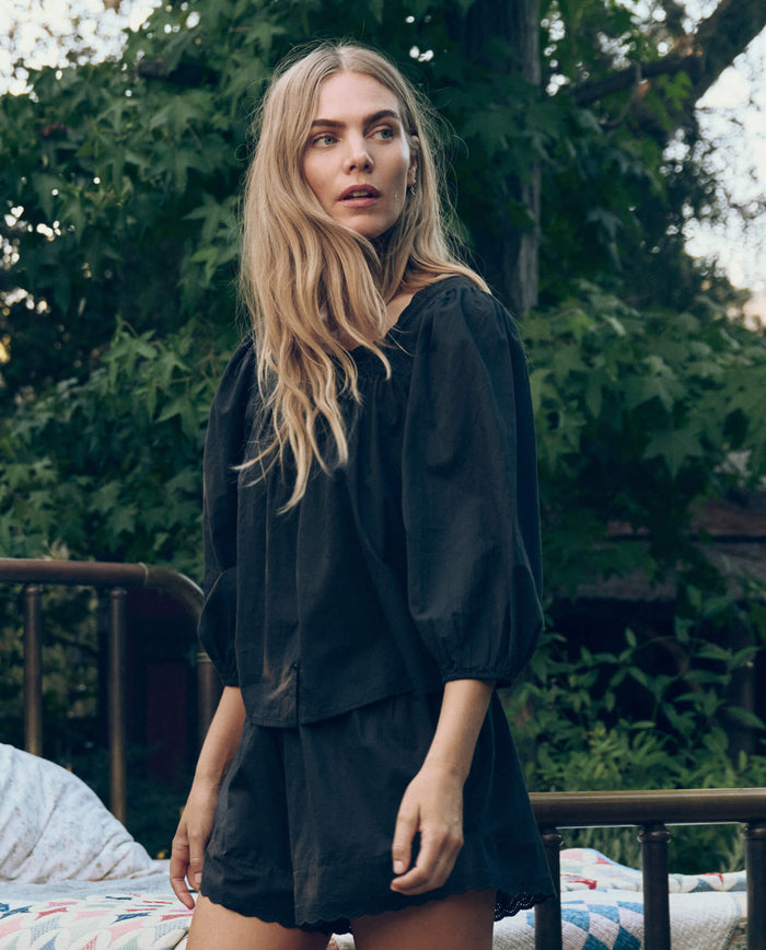 A woman with long blonde hair stands outdoors in front of green foliage, wearing THE GREAT. CORE SLEEP's The Eyelet Button Sleep Top in black with Victorian-inspired lace, paired with matching shorts.
