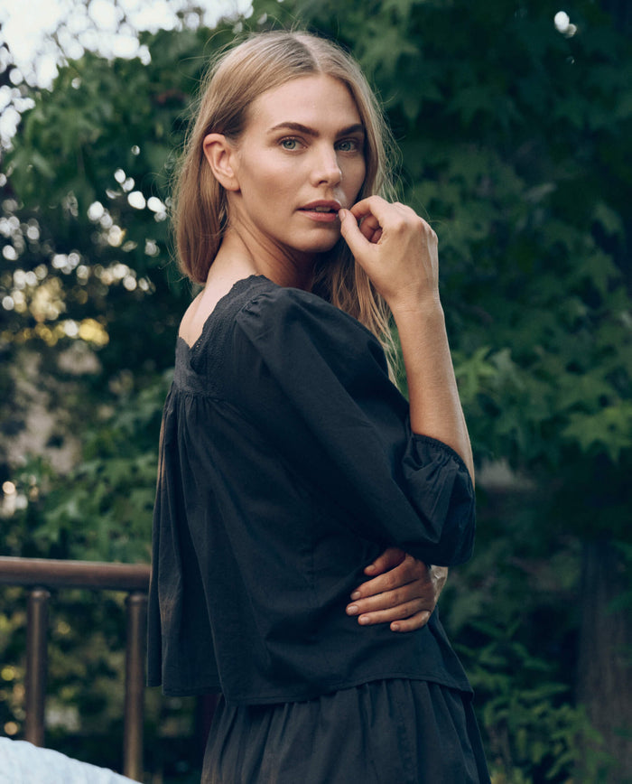 A woman with long blonde hair stands outdoors amid green foliage, wearing THE GREAT. CORE SLEEP's The Eyelet Button Sleep Top in Black and looking at the camera with her hand near her mouth.