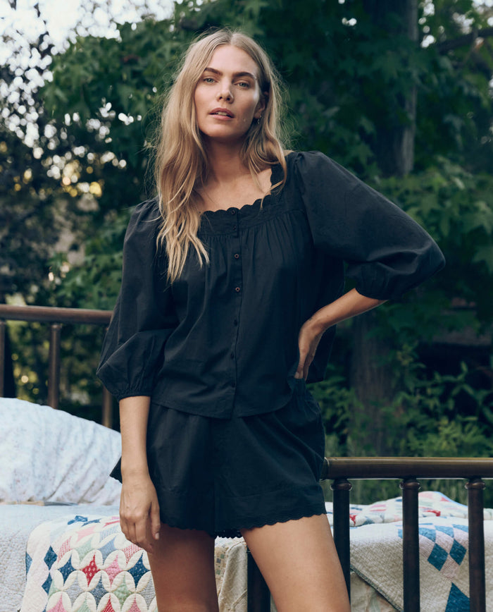 A woman with long blonde hair stands outdoors in THE GREAT. CORE SLEEP’s The Eyelet Button Sleep Top in black. A metal bed with a colorful quilt appears in the background among the trees.