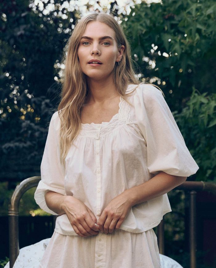 A woman with long blonde hair stands outdoors amid green foliage, wearing THE GREAT. CORE SLEEP's The Eyelet Button Sleep Top in True White with Victorian lace trim and buttons.