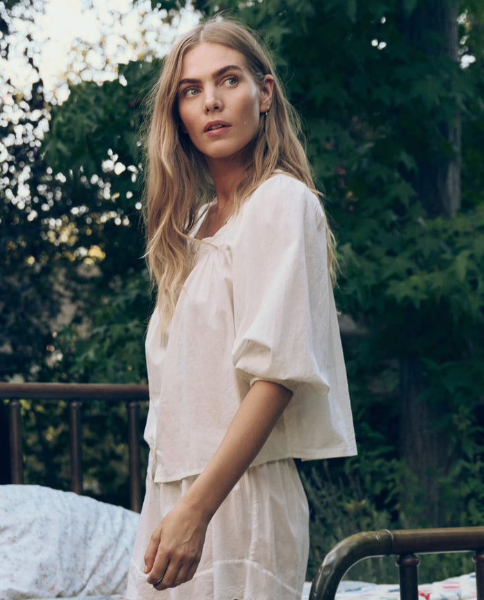 A woman with long blonde hair stands outdoors beside a metal bed frame, wearing THE GREAT. CORE SLEEP's Eyelet Button Sleep Top in True White, featuring delicate Victorian lace trim, with trees in the background.