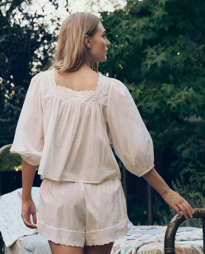 Wearing THE GREAT. CORE SLEEP’s Eyelet Button Sleep Top in True White, a blonde-haired person stands outdoors by a metal bed frame, exuding effortless comfort in the loose-fit, long-sleeved cotton top and matching shorts.